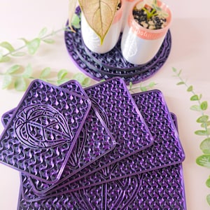 May include: A stack of purple, textured coasters with a leaf design. The coasters are square with a repeating pattern and a central leaf shape. In the background, small potted plants sit on matching purple saucers.