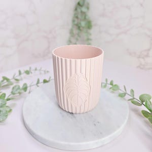 May include: A pale pink cylindrical planter featuring a vertical ribbed texture on the exterior surface. The front of the planter displays a raised, embossed leaf design centered within the vertical grooves, creating a consistent pattern.