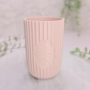 May include: A pale pink cylindrical planter featuring a vertical ribbed texture on the exterior and a debossed leaf pattern centered on the front. The vessel has an open top and a rounded base, providing a structured container for small plants.