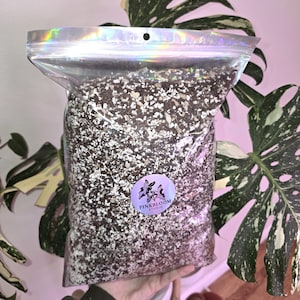 May include: A clear, holographic zip-lock bag filled with a soil mixture. The soil is a mix of dark brown, white, and tan particles. A round sticker with the text "PINKBLOOM STUDIO" and a floral design is on the bag. The bag is held in front of a plant.