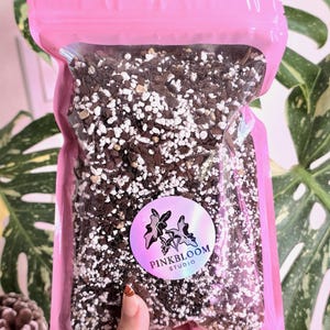 May include: A pink, resealable bag filled with a soil mix, featuring a mix of dark brown, white, and light brown components. A circular sticker with the text "PINKBLOOM STUDIO" and a floral design is visible on the bag.