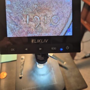 May include: A black Elikliv digital microscope with a rectangular screen displaying a magnified view of a copper-colored surface with the embossed date 1910. The device features control buttons labeled OK, left arrow, right arrow, MENU, and a power icon.