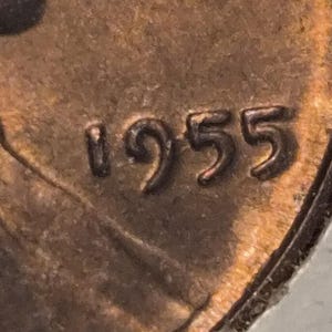 Könnte beinhalten: Eine Nahaufnahme einer kupferfarbenen Münze mit der eingeprägten Jahreszahl 1955 auf der Oberfläche. Das Metall weist eine verwitterte, strukturierte Patina mit sichtbaren Anzeichen von Alterung und Oxidation auf der runden Münzfläche auf.