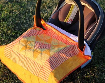 May include: A gray infant car seat with a black handle and a warning label, covered by a quilted blanket featuring a geometric star pattern in shades of orange, yellow, and white with striped and dotted fabric borders and a solid orange edge.