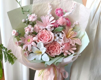 May include: A handmade bouquet of crochet flowers in pink, white, and green. The bouquet features roses, lilies, and other floral designs, wrapped in tissue paper and tied with a pink ribbon. The flowers are held by a person.