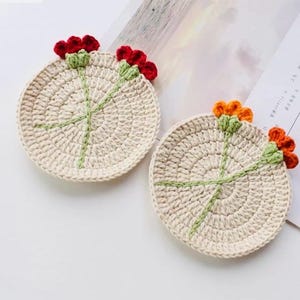 Puede incluir: Dos posavasos de ganchillo hechos a mano. Cada posavasos es circular, con una base de color crema y un diseño floral. Las flores son rojas y naranjas, con tallos y hojas verdes. Los posavasos están sobre una superficie blanca.