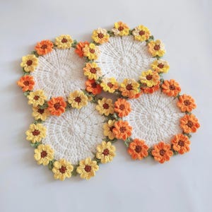 Puede incluir: Cuatro posavasos de ganchillo hechos a mano. Cada posavasos es blanco con un diseño circular y un borde de flores de ganchillo amarillas y naranjas con hojas verdes. Los posavasos están dispuestos sobre una superficie blanca.
