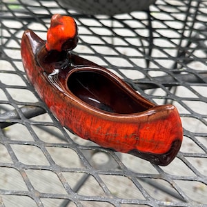 Puede incluir: Una jardinera de cerámica con forma de canoa, con un esmalte brillante moteado rojo y marrón. La jardinera presenta una pequeña figura redondeada en la popa. El interior es marrón oscuro y la jardinera está sobre una rejilla metálica.