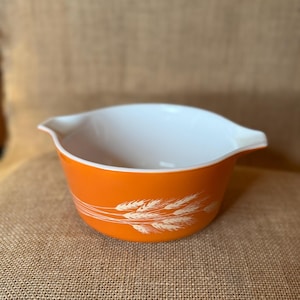 May include: An orange Pyrex bowl with a white interior and two handles. The exterior features a wheat pattern in white. The bowl is set against a neutral background.
