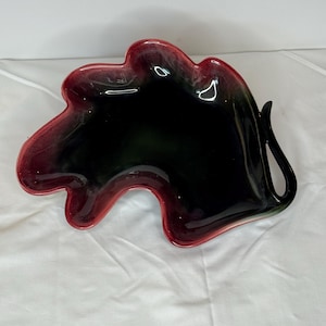 May include: A leaf-shaped decorative dish with a glossy finish. The dish features a gradient of colors, with a deep red rim that fades into black and green in the center. The dish is made of ceramic or glass.