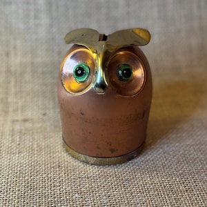May include: A copper-colored owl-shaped bank with brass accents. The owl has green eyes and a slot on top for inserting coins. The bank is made of metal and has a vintage appearance.