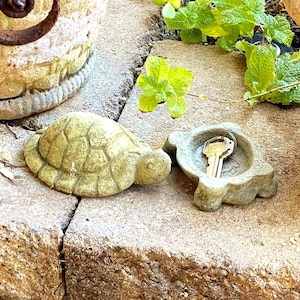 May include: A two-piece stone turtle figurine with a textured, light green finish. The turtle consists of a solid shell top that lifts to reveal a hollowed-out base, which currently holds a single silver-colored metal key inside its compartment.