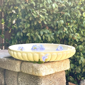 May include: A shallow, round, off-white stone birdbath basin featuring a scalloped, fluted exterior edge. The basin is filled with clear water and contains several small, light blue flowers floating on the surface of the water.