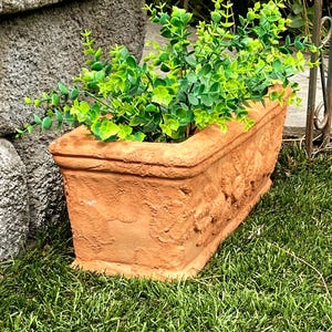 May include: A rectangular terracotta planter filled with vibrant green foliage. The planter has a textured surface with embossed floral designs. It sits on a bed of green grass, with a stone wall and metal fence in the background.