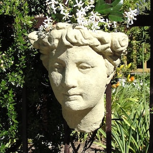 May include: A beige stone-colored planter shaped like a human head with a textured surface. The sculpture features a crown of hair and a hollow top filled with small white star-shaped flowers and green leaves, mounted on a dark metal fence.