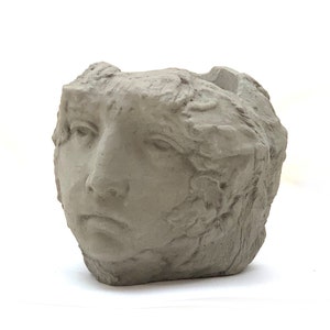 May include: A light gray concrete planter shaped like a human head. The vessel features a textured, rough surface with detailed facial features including eyes, a nose, and lips. The top of the head is open, creating a hollow space for plants.