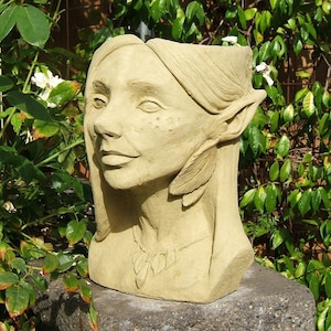 May include: A beige ceramic planter shaped like a woman's head with long hair and an ear that is pointed like an elf's ear. The planter is sitting on a gray stone surface.
