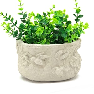May include: A small, round, off-white ceramic planter with embossed floral and bird designs. The planter is filled with vibrant green artificial eucalyptus plants, creating a fresh, decorative accent. The pot is approximately 5 inches in diameter.