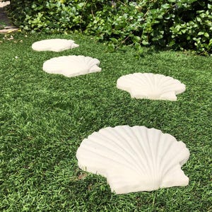 May include: A set of four off-white, seashell-shaped stepping stones arranged on a bed of green grass. The stones have a textured surface with detailed fan-like ridges. The stones are a decorative garden element.