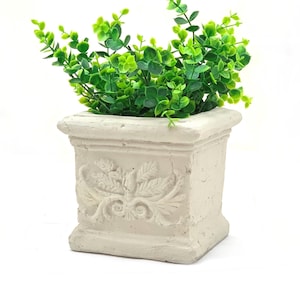 May include: A square, off-white planter with a detailed, embossed floral design. The planter holds vibrant green faux eucalyptus. The planter has a classic, aged appearance, adding a touch of elegance to any space. The overall aesthetic is a blend of rustic and refined.