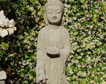 May include: A light gray stone statue of a standing Buddha figure with closed eyes and a serene expression. The figure wears traditional robes with detailed trim and holds a small lotus flower in both hands, positioned at the center of the chest.