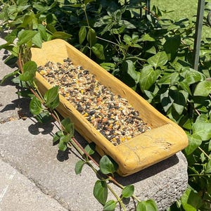 May include: A rustic, rectangular wooden bird feeder filled with birdseed. The feeder is a light yellow color and sits on a gray stone surface, surrounded by green foliage. The birdseed is a mix of grains and seeds.