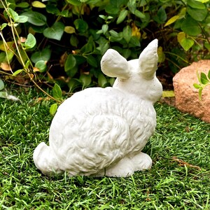 May include: A white, textured rabbit figurine sculpted in a seated position with upright ears. The surface of the rabbit features detailed fur-like carvings, capturing the animal's form in a static pose, suitable for outdoor or garden display.