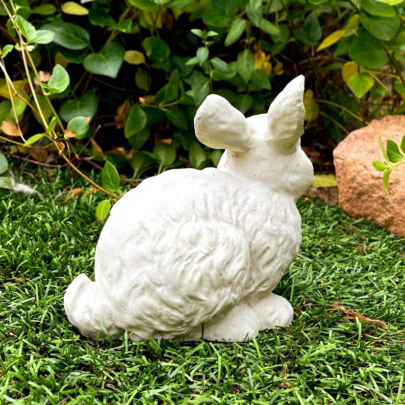 May include: A white, textured rabbit figurine sculpted in a seated position with upright ears. The surface of the rabbit features detailed fur-like carvings, capturing the animal's form in a static pose, suitable for outdoor or garden display.
