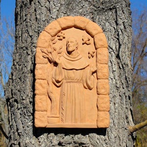 May include: A terracotta relief sculpture of a religious figure within an arched frame. The figure is depicted with arms outstretched, wearing a robe. The frame has a brick-like texture, and the background shows a tree trunk.