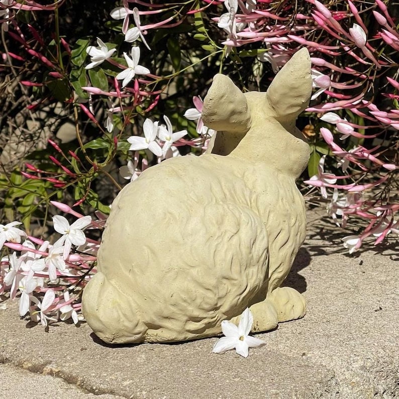 May include: A light beige, textured rabbit figurine in a seated position, featuring sculpted fur details, upright ears, and a rounded tail, positioned against a backdrop of green foliage with small white and pink star-shaped jasmine flowers.