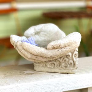 May include: A light beige, stone-like decorative bowl shaped as two cupped hands joined together. The exterior base features an intricate, raised scrollwork pattern, while the interior of the hands holds a small, light purple fabric petal.
