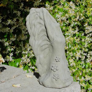 May include: A light gray concrete garden sculpture featuring a vertical, abstract form with a textured, wavy surface on one side and a smooth, flat side adorned with three small, raised flower carvings and the embossed text TRIBUTE at the base.