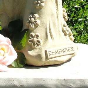 May include: A cream-colored stone sculpture featuring a base adorned with sculpted floral motifs and the word REMEMBER embossed in a rectangular plaque. A pale pink rose with green leaves is positioned to the left of the textured stone base.