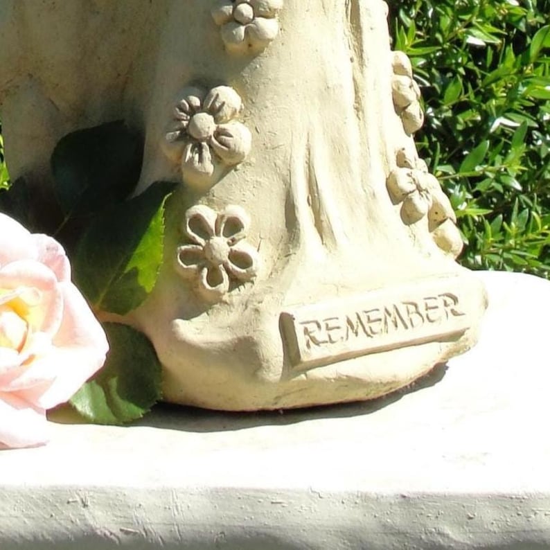 May include: A cream-colored stone sculpture featuring a base adorned with sculpted floral motifs and the word REMEMBER embossed in a rectangular plaque. A pale pink rose with green leaves is positioned to the left of the textured stone base.