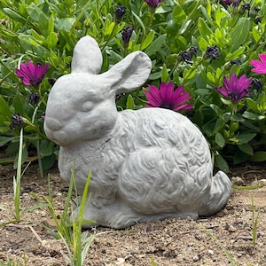 May include: A light gray concrete rabbit figurine in a seated position, featuring textured fur details, long upright ears, and a rounded tail, positioned on the ground in front of a patch of green foliage with vibrant purple flowers.