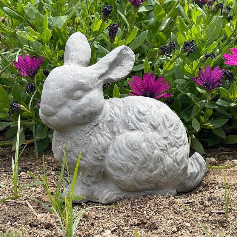 May include: A light gray concrete rabbit figurine in a seated position, featuring textured fur details, long upright ears, and a rounded tail, positioned on the ground in front of a patch of green foliage with vibrant purple flowers.