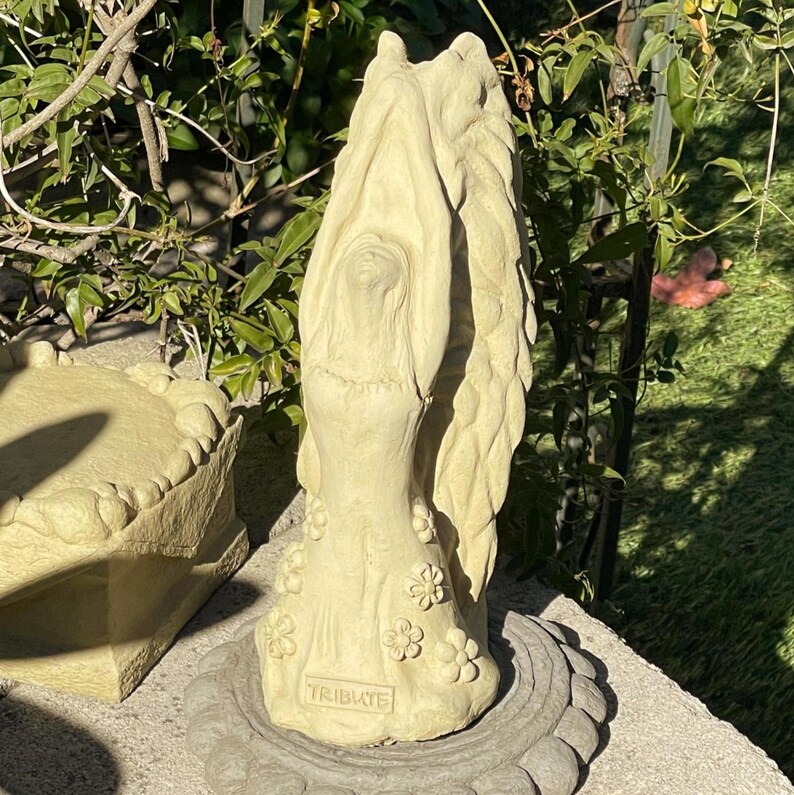 May include: A light beige stone sculpture of a figure with arms raised above their head, featuring large wings on the back and floral details on the dress. The base of the sculpture has the word TRIBUTE embossed in a rectangular frame.