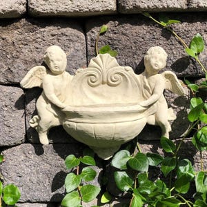 May include: A beige-colored wall planter featuring two cherubic figures with wings, holding a decorative urn-shaped container. The planter is mounted on a textured stone wall, with green ivy vines adding a natural touch.
