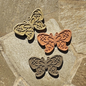 May include: Three butterfly-shaped wall ornaments made of molded material, each featuring an intricate raised scrollwork and floral pattern. The set includes one light beige, one terracotta orange, and one dark gray butterfly, all with a matte finish.
