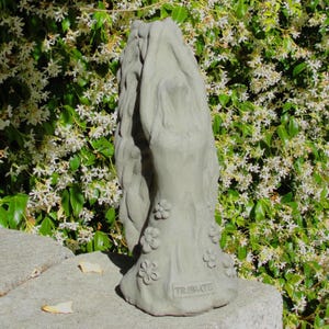 May include: A light gray stone sculpture depicting a figure with long flowing hair reaching upward, adorned with small sculpted flowers around the base. The word TRIBUTE is embossed in capital letters on the lower front of the statue.