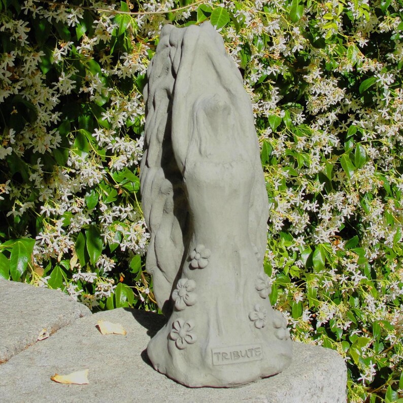 May include: A light gray stone sculpture depicting a figure with long flowing hair reaching upward, adorned with small sculpted flowers around the base. The word TRIBUTE is embossed in capital letters on the lower front of the statue.