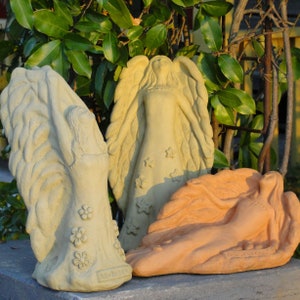 May include: Three stone-like angel sculptures. Two are light beige, one standing with flower details and one standing with star details. The third is a terracotta-colored reclining angel sculpture. Each figure features large, textured wings and flowing robes.
