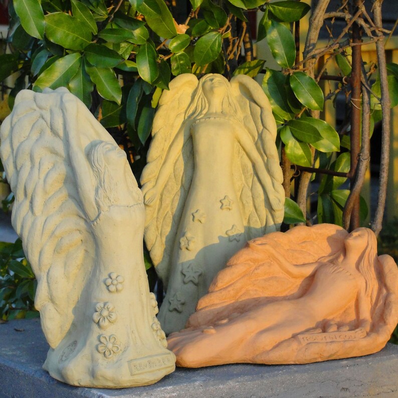May include: Three stone-like angel sculptures. Two are light beige, one standing with flower details and one standing with star details. The third is a terracotta-colored reclining angel sculpture. Each figure features large, textured wings and flowing robes.