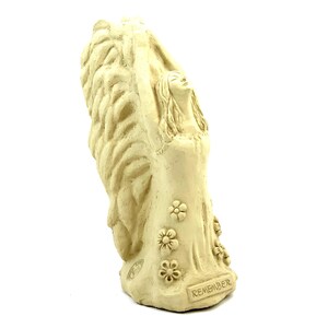 May include: A cream-colored stone-like figurine of a figure with long hair and large, textured wings reaching upward. The figure wears a dress adorned with embossed flower patterns and features the word REMEMBER engraved on the base.