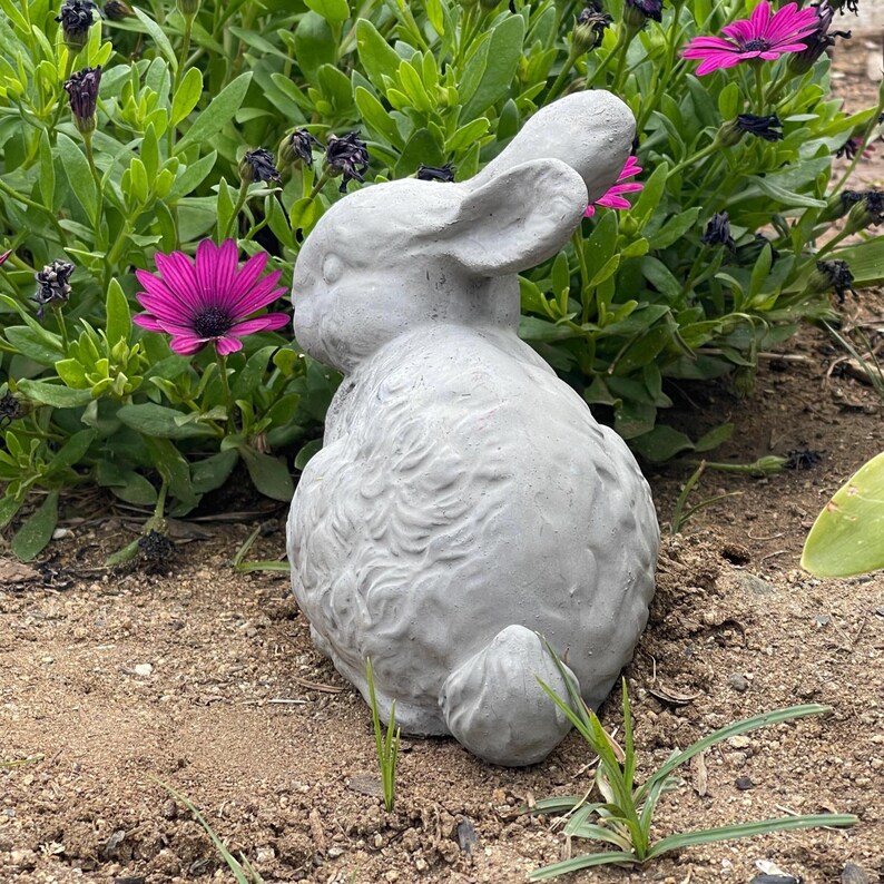 May include: A gray, textured rabbit figurine in a seated position, featuring detailed fur-like carvings, long ears, and a small tail. The sculpture is positioned on a patch of dirt, showcasing a realistic form and a neutral, matte finish.