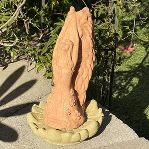 May include: A terracotta-colored garden statue of a figure with wings reaching upward, standing on a light green, leaf-shaped base. The figure wears a dress adorned with small flower carvings and features the word REMEMBER embossed at the base.