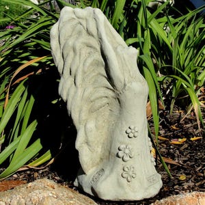 May include: A light gray stone garden statue depicting a figure with long, flowing hair and arms raised upward. The figure wears a dress adorned with three-dimensional flower embellishments along the side and a circular emblem near the base.