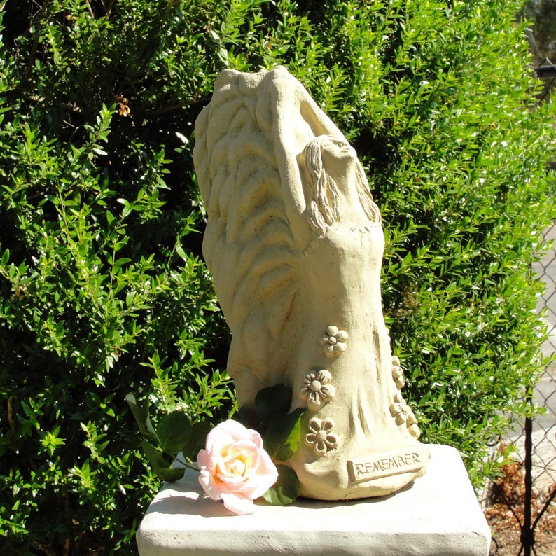May include: A beige stone-colored sculpture of a figure with large wings reaching upward, adorned with small sculpted flowers and the word REMEMBER embossed at the base, accompanied by a single light pink rose with green leaves at the bottom.