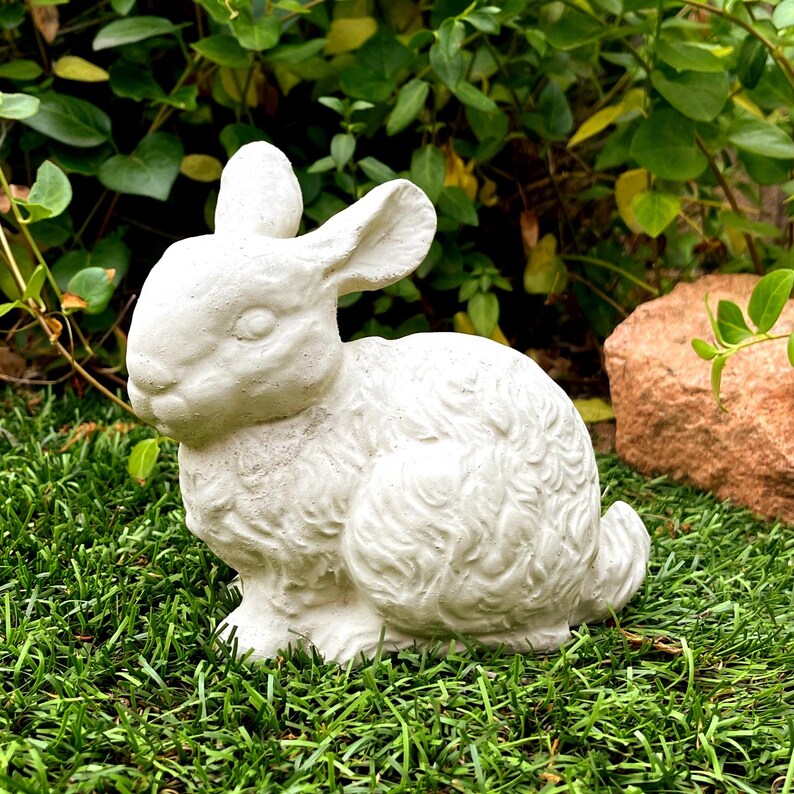May include: A white, textured concrete rabbit figurine in a seated position. The sculpture features detailed fur patterns, long upright ears, and a rounded body, capturing the likeness of a small animal with a matte, off-white finish.