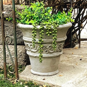 May include: A light gray concrete planter with a flared rim and a decorative embossed scroll pattern around the center. The planter is filled with vibrant green artificial eucalyptus foliage and trailing string of pearls succulent stems.